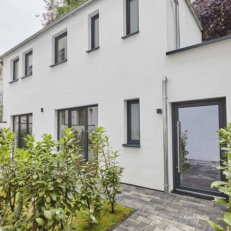 Remise Fassade - Weiße Fassade eines zweistöckigen Gebäudes mit mehreren Fenstern, Glastür, davor ein kleiner Garten mit Hecke und gepflastertem Weg