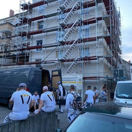 Mehrere Personen in weißer Arbeitskleidung sitzen und stehen vor einem mehrstöckigen Gebäude mit Gerüst und Treppenanlage, zwei Fahrzeuge sind geparkt.