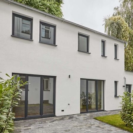 Remise Fassade - Weiße Fassade eines zweistöckigen Gebäudes mit mehreren Fenstern, Glastür, davor ein kleiner Garten mit Hecke und gepflastertem Weg