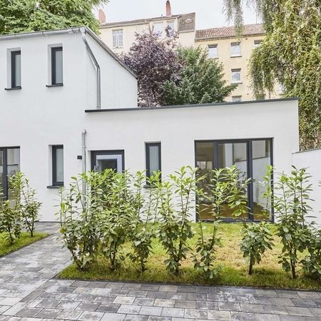 Remise Fassade - Weiße Fassade eines zweistöckigen Gebäudes mit mehreren Fenstern, Glastür, davor ein kleiner Garten mit Hecke und gepflastertem Weg