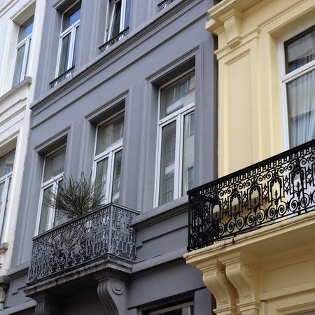 Fassaden von drei mehrstöckigen Gebäuden mit Fenstern und dekorativen Balkonen, einer grau, einer gelb und einer weiß gestrichen.